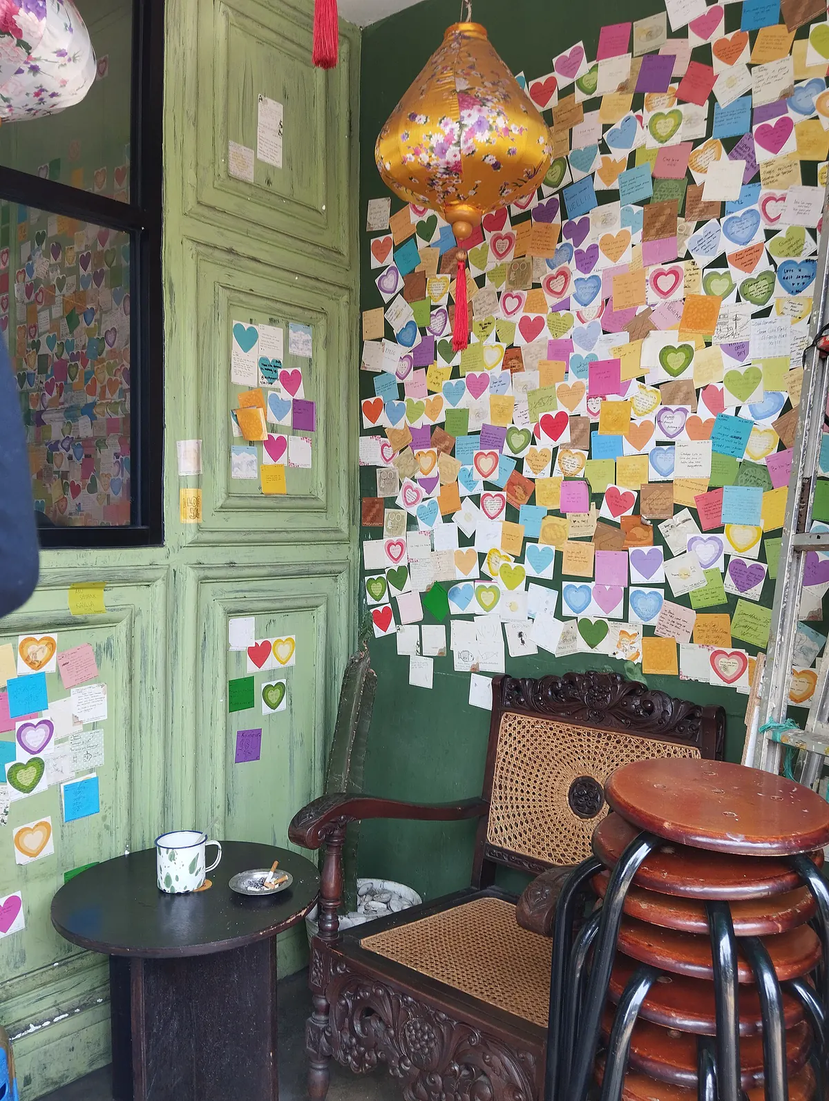 Sudut kafe di Kampung Kayutangan dengan pintu hijau tua, dinding catatan warna-warni, dan kursi rotan
