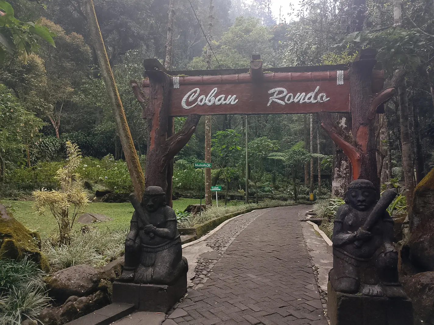 Gerbang kayu bertuliskan Coban Rondo dengan dua arca penjaga di awal jalur paving