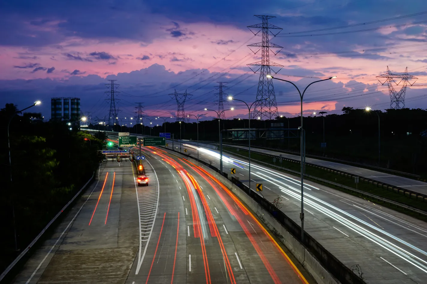 Jalan tol saat senja dengan jejak lampu kendaraan dan menara listrik di bawah langit ungu