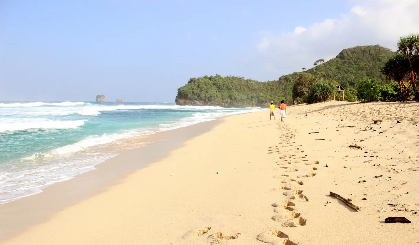 Dua pengunjung berjalan di pasir putih yang landai menuju bukit hijau di tepi laut